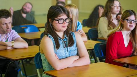 A girl with dark hair and arms crossed sitting at a school desk, alongside other pupils also sitting at desks in a classroom. 