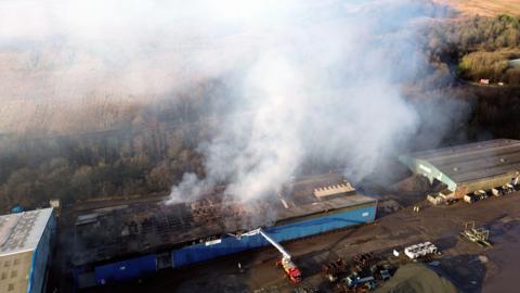 A drone image of the industrial estate which shows the wrecked buildings and billows of smoke above them. There are some vehicles on the site, including a fire engine, and trees and fields in the background.