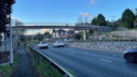 A two lane road with cars driving away from the camera, with a grey footbridge above the road and a blue sky with clouds in the background.