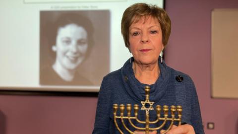 A woman holding a traditional Jewish candle, a hanukkiah, is standing in front of a large screen mounted on the wall, showing an image of her mother behind.