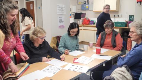 The image shows people gathered around a table, taking part in art therapy provided by the charity Arty Folk. They are all women and, on the table, are several pieces of paper with writing and images on them. Behind the table is a small kitchen with cupboards.