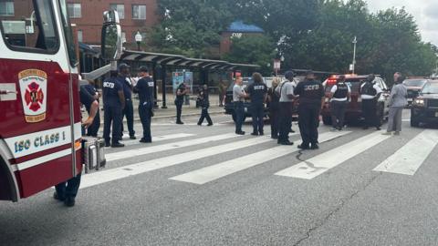 A landscape image of a road crossing with multiple emergency service workers standing around. The front of a red fire truck can be seen on the left hand side.