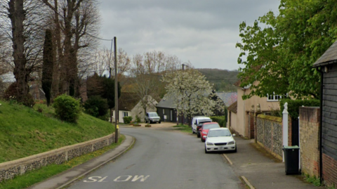 A Google Streetview image of Ickledon Road in Elmdon. The grass bank beside a church is visible on the right, and there are cars parked on the pavement to the right, as well as houses and trees.