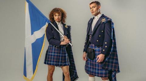 A male and female model in the Team Scotland uniform - blue tartan kilts and blue jackets. The female model is holding a saltire over her shoulders