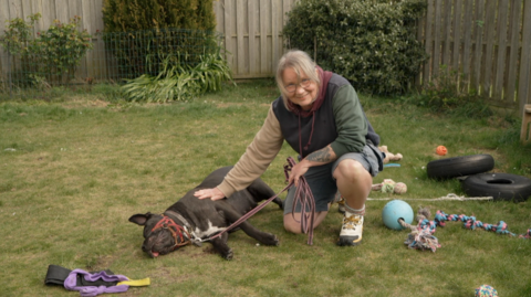 Nina Morgan kneels on the grass in a garden, gently resting a hand on a large black dog named Frankie lying on its side. Frankie is wearing a red head collar and leash. Various dog toys, including ropes and balls, are scattered around the garden, which is enclosed by wooden fencing and greenery.