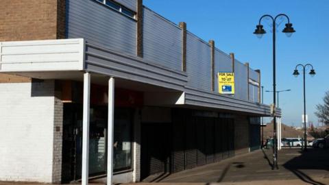 A view of Eston Precinct. Metal shutters are down across a number of properties and a yellow For Sale or To Let sign is attached to one part of the building.