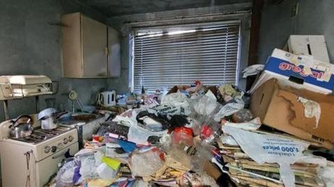 A kitchen piled high with rubbish including plastic bags, newspapers and cardboard boxes. In the background is a small hob, some shelving and a kettle. 