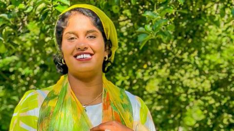 A woman with brown hair wears a green chiffon top and hair scarf. She is smiling outside in a garden.