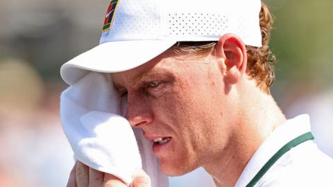 Jannik Sinner, in white tennis shirt with green trim and white cap, mops away sweat from his face