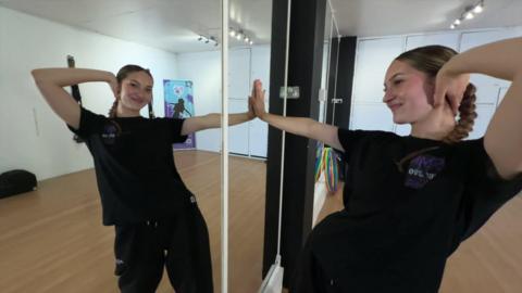 Maria Radu wearing all black with her hair tied back is seen posing in a mirror that is located in her dance studio. She has one hand on her head and another flat against the mirror. 