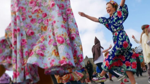 A woman in a patterned, blue dress and wearing a flower garland dances outside. In front of her is a close-up of a light blue, floral patterned dress and behind her are people also dancing