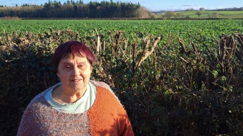A woman with short dark red hair is stood next to a large hedge. She is wearing a blue t-shirt and an orange jumper.