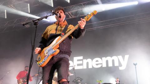 The Enemy performing at Godiva Festival with the band's front singer at a microphone while holding a guitar while behind him is a backdrop with "The Enemy" written on it