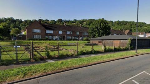 A google street view screenshot as captured from across the road. It is a large grassy patch with a fence around it, protecting the remains of a Roman villa. On the right there is a large wooden single storey shed. Behind the remains are a row of brick houses and trees.