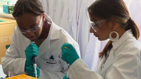 Two teenage women wearing white lab coats, lab glasses and green rubber gloves, using pipettes.