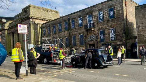 Film crew members gathered around a truck, equipped with a camera rig, in front of a classic car. A large theatre building can be seen on the opposite side of the road.