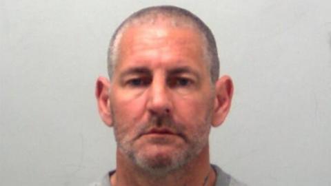 A custody photo of Mitchell Rudkin. He is wearing a grey jumper and has a shaved head.