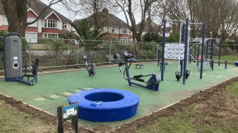 An outdoor gym area in a park. There are different types of gym equipment on a fenced off portion of AstroTurf. 