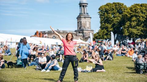 Beth Heath has shoulder-length brown hair and is wearing a pink t-shirt and black trousers. She has both arms out in the air. She is standing on green grass with dozens of people sitting and standing on the grass behind her. In the background is a church and white tents.