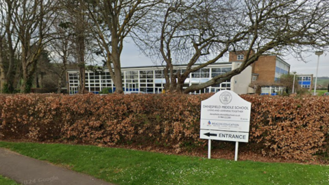 A view of a school building. The sign in front says Danesfield Middle School. The hedge is brown and the trees have no leaves.