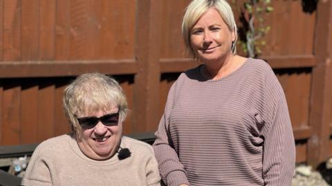 Resident Linda sitting down with cropped white/blonde hair and sunglasses while care support worker Donna stands next to her wearing a purple long-sleeved top and a blonde bob