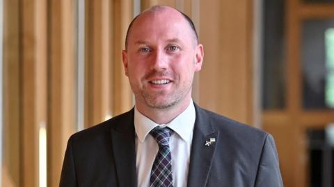 Neil Gray, a bald man with a grey suit, smiles as he walks in the Scottish Parliament. He is photographed in a medium close-up. 