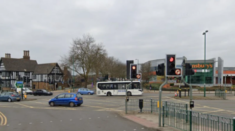 A road junction with a number of cars and a white bus on it and traffic lights and metal railings in the foreground. There is a Sainsbury's store and a black and white timbered building in the background