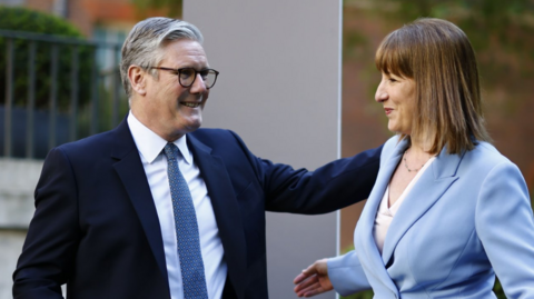 Sir Keir Starmer pats Chancellor Rachel Reeves on the shoulder. She wears a powder blue suit while he is in a darker navy number. 