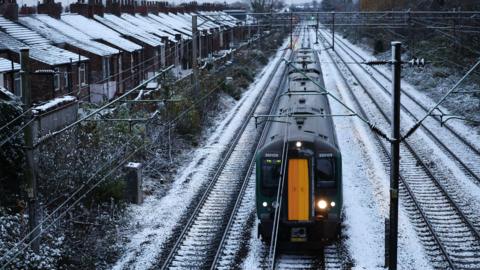 Library photograph of a black and yellow train as it travels on tracks covered in snow. Houses on the left of the tracks have snow on their roofs.