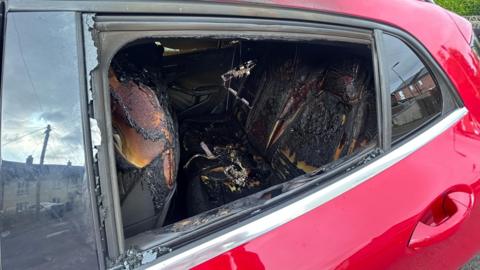 A red car with the back passenger window smashed. The interiors are burnt.