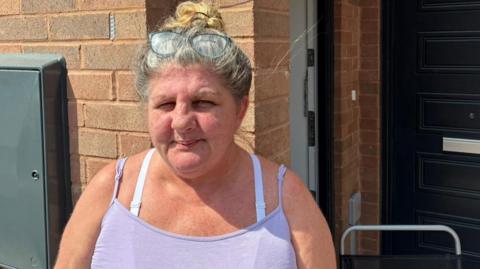 Debbie has her hair tied back in a bun and is wearing a purple vest. She is stood outside her front door.