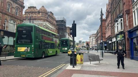 A street in Leeds with police tape across the road, two green double decker buses are parked up and there are several police cars in view. 