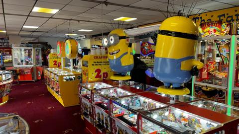 An amusement arcade. Slot machines and 2p change machines are visible. Two minions figures from the Despicable Me films sit atop the slot machines.