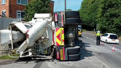An overturned cement lorry has crashed into a brick wall and fence. Police officers are stopping traffic in the road next to it. The lorry is white with black tyres and red and yellow "highway maintenance" markings on the back. A house and trees can be seen to either side of the road.