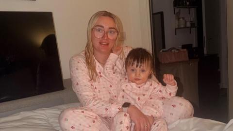 Maisy May Webb and her two-year-old daughter are pictured sitting on a bed. They are wearing matching pink and white pyjamas and both appear to have wet hair. There is a large TV in the background.