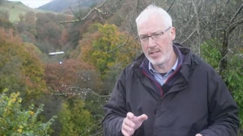 BBC Reporter Craig Duggan stands in a valley of autumn trees, the train carriage of one of the crashed trains is visible in the background. Craig Duggan is wearing a black raincoat and has grey hair and is wearing glasses. 