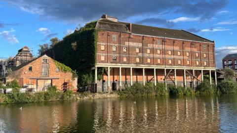 Downing's Malthouse at Baker's Quay in Gloucester. It is a large red brick building right on the waterfront, with pillars supporting the upper levels. It is very overgrown, with boarded up windows and fading signage.