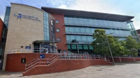 The orange brick and glass building has stairs leading to it on the left and right, in front of it. There are some tall tree on the right shielding some of the glass near to some lamp posts.