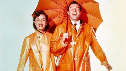 A man and a woman in orange rain jackets with bright faces smiling with their mouths open. The man is wearing a suit underneath his jacket and holding an orange umbrella over the two of them.