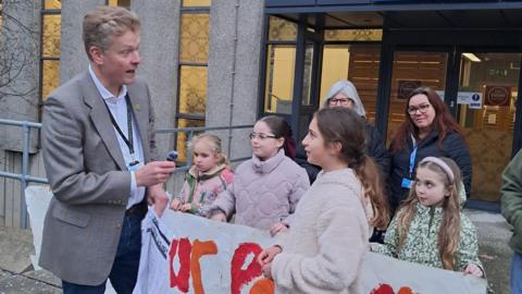 Lottie and friends stand outside the Town Hall. A blond-haired man, Matthew Snedker, in a blazer and jeans, faces Lottie holding a small microphone. Lottie has long brown hair, tied back in a pony tail, and is wearing a cream jumper. Behind them, a small group of young girls hold up a homemade painted banner.
