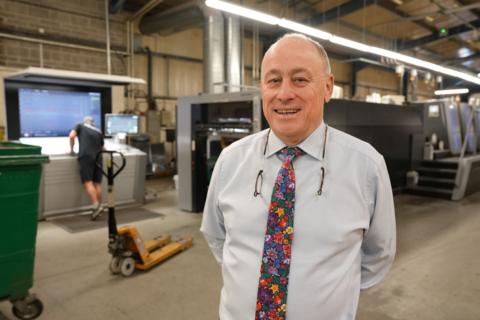 A man in a shirt and floral tie stands in front of printing machinery