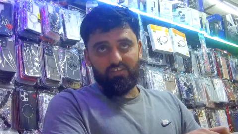 Shop worker inside a mobile phone accessories shop, behind a glass counter, with walls densely covered in packaged phone cases, headphones and electronic accessories.