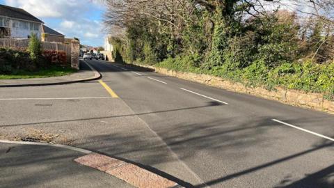 La Grande Route De St Clement at the entrance of Clos Des Patelles - a main road with an estate of houses on the left and branches and trees on the right. Cars in the distance.