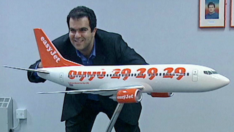 Stelios Haji-Ioannou, wearing a suit stood in front of a model EasyJet plane, and smiling for a picture in an office.