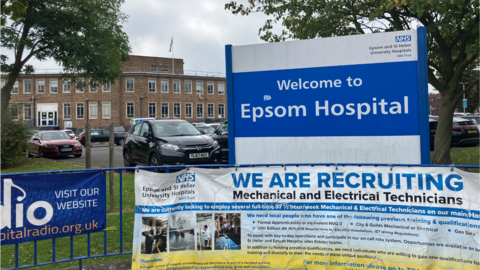 Sign outside Epsom hospital, fence with recruitment advert, cars, hospital building