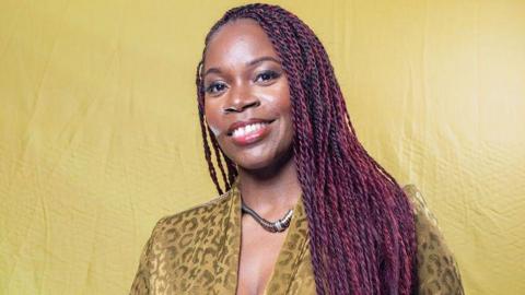 Jacqueline Shaw has long, dark purple braided hair. She is wearing a necklace and a light khaki patterned coloured top. She is standing in front of a light khaki background.