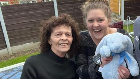 An older woman, two young women and a young man stand smiling at the camera as the young woman on the right takes a "selfie". The other young woman is holding a blue stuffed rabbit. 