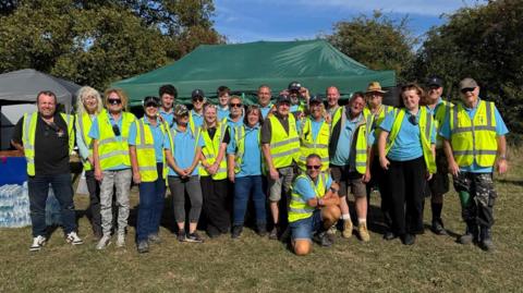 A large group of people wearing high-vis vests are gathered for a photo. They are stood in front of a green gazebo in a field. 