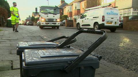 Two black plastic food caddies - about knee high - sit on a pavement in a Newcastle suburb. A council waste worker clad in a high-vis t-shirt and shorts strolls towards them in the background alongside a white waste removal van with headlights shining forward in the morning sun. 