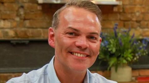 MasterChef winner Simon Wood smiles for the camera wearing a blue shirt with a vase of flowers seen behind him on a table in front of a brick wall. 